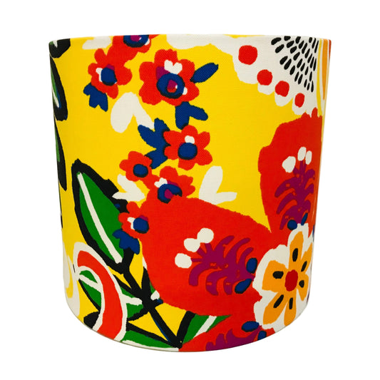 A cylindrical lampshade featuring a vibrant retro floral pattern on a bright yellow background, with red, orange, blue, green, and white motifs.