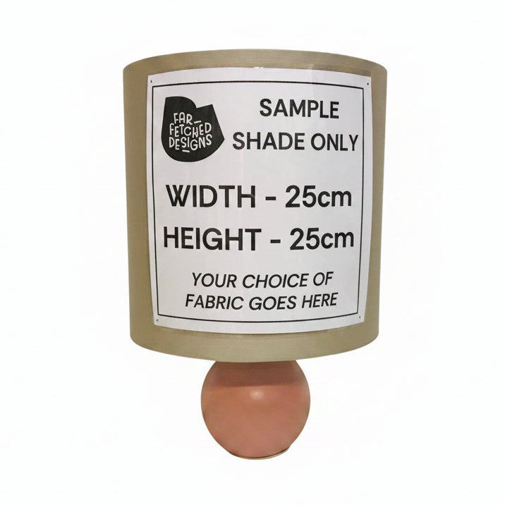 A cylindrical lampshade sample with a printed label reading 'SAMPLE SHADE ONLY' and dimensions 'WIDTH - 25cm HEIGHT - 25cm', mounted on a pink round base against a plain off-white wall.