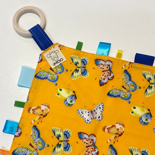 A colorful baby taggie toy with a variety of fabric tags and a wooden ring, backed with an organic bamboo toweling.