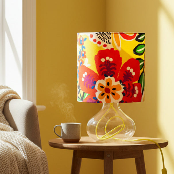 A table lamp with a vibrant, multicolored floral patterned drum shade and a clear glass base with a yellow cord