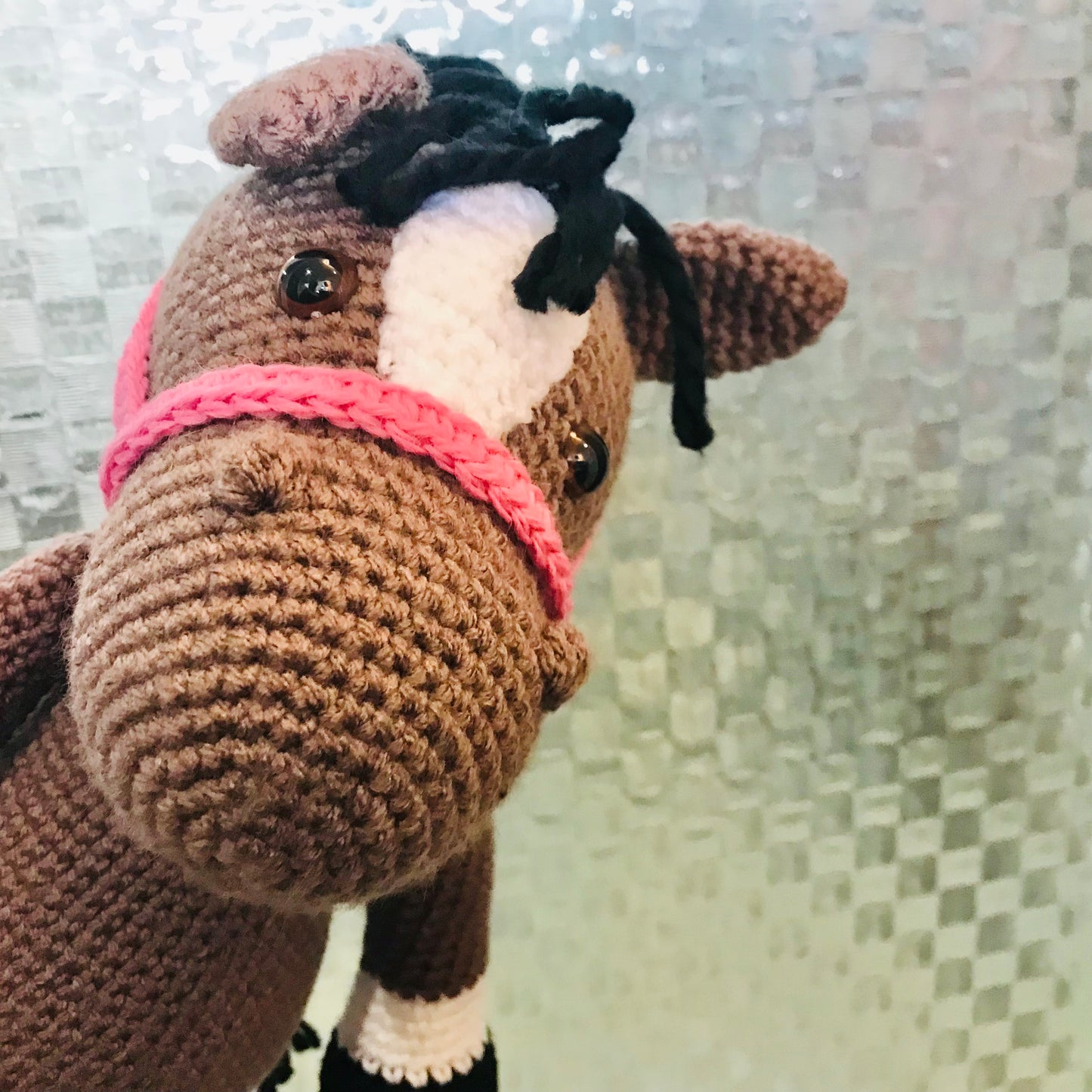 Crocheted horse toy with a pink bridle against a textured wall.