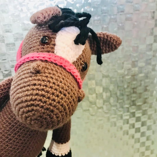 Crocheted horse toy with a pink bridle against a textured wall.