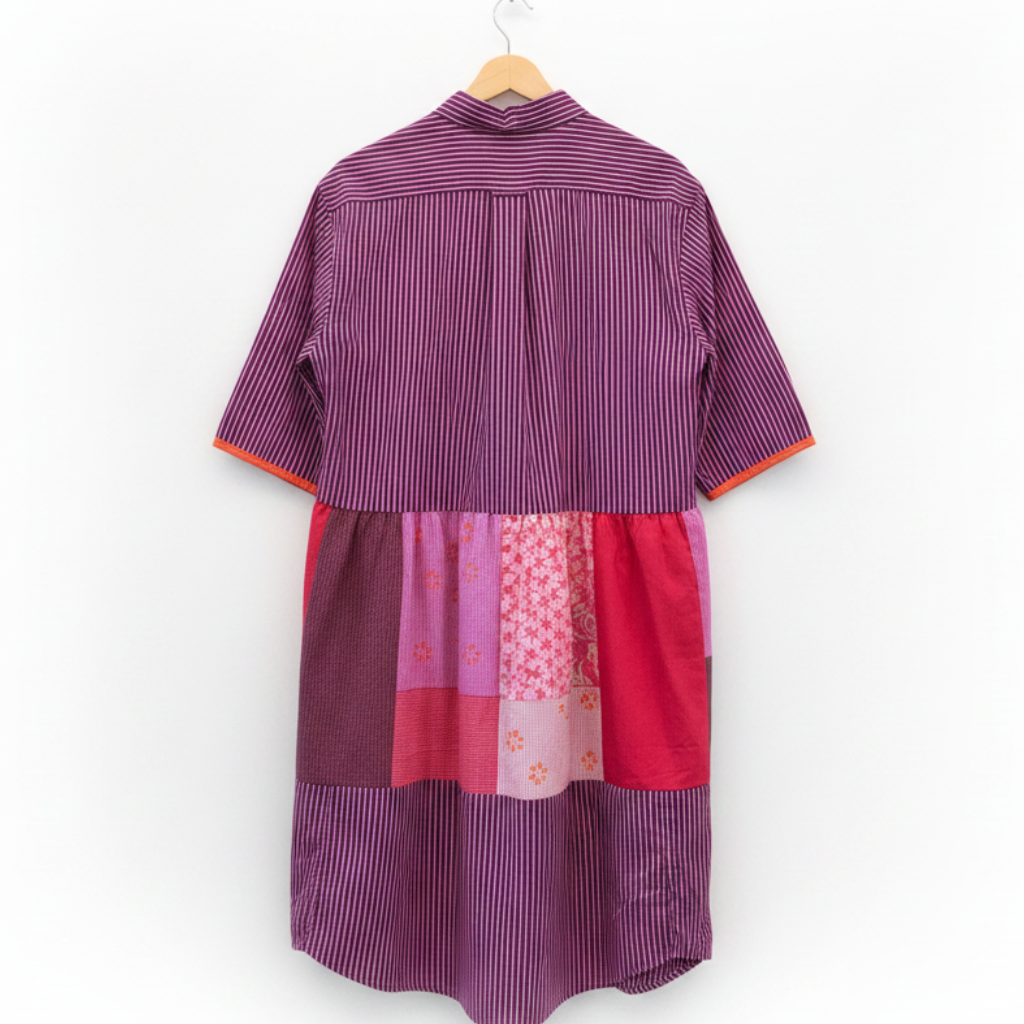A purple and pink striped patchwork shirt dress hanging on a wooden hanger against a white brick wall.