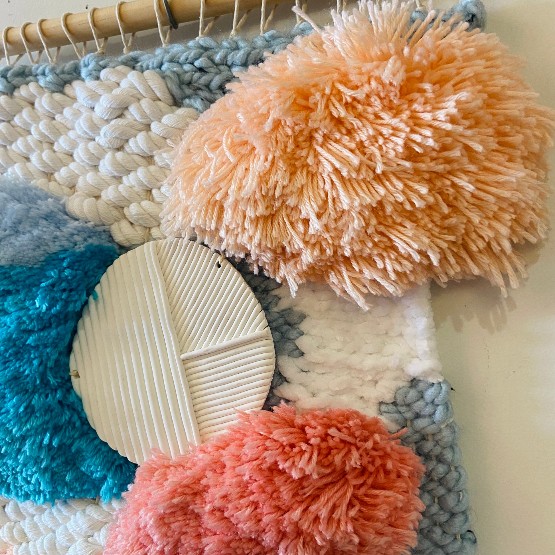 Textured wall art with colorful yarn and pom-poms on a wooden hanger.
