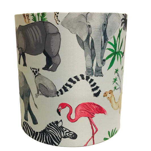 A cylindrical lampshade featuring a safari-themed animal print with a white background and illustrations of a pink flamingo, zebra, elephant, rhino, lemur, camel, and green palm leaves.