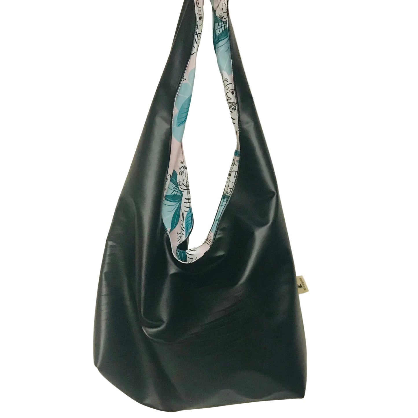 A black faux-leather slouchy shoulder bag with a colorful tropical leaf print fabric lining the strap, hanging against a plain off-white wall.