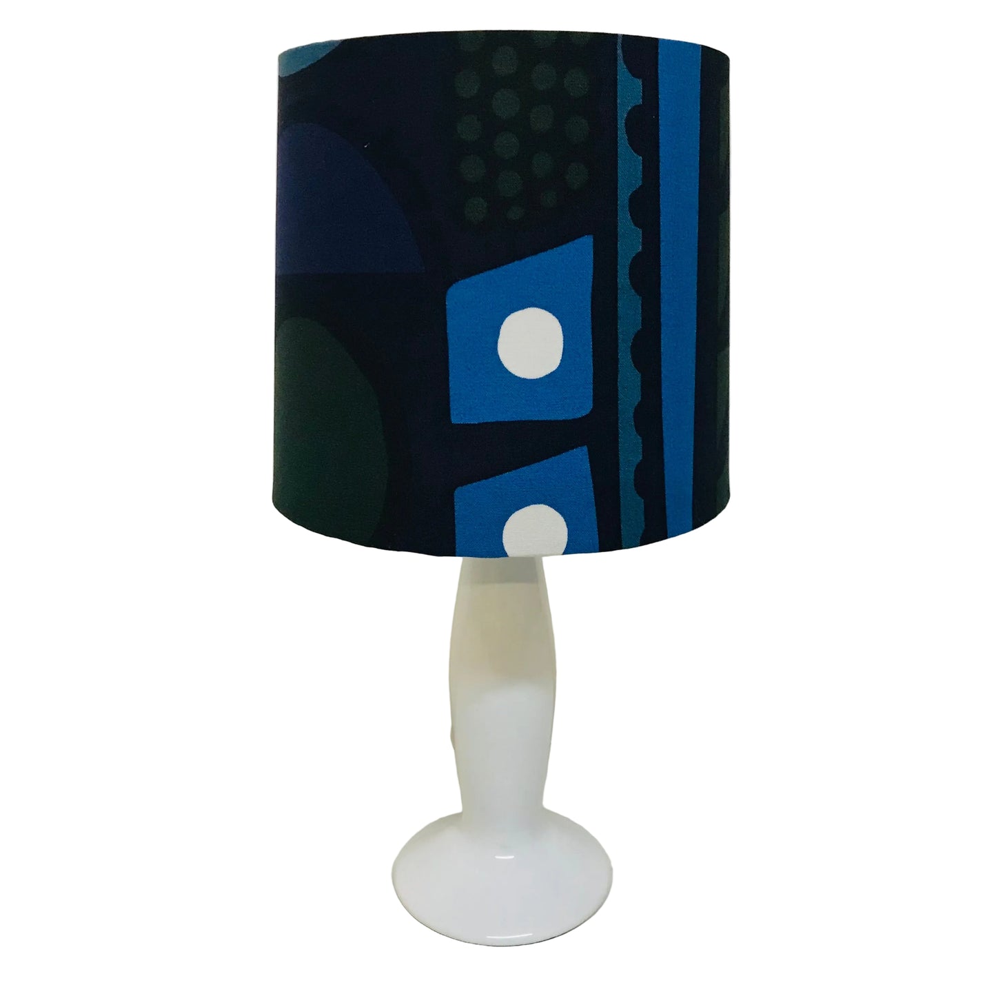 A table lamp with a cylindrical drum shade featuring a blue, black, and white geometric pattern, mounted on a glossy white pedestal base.
