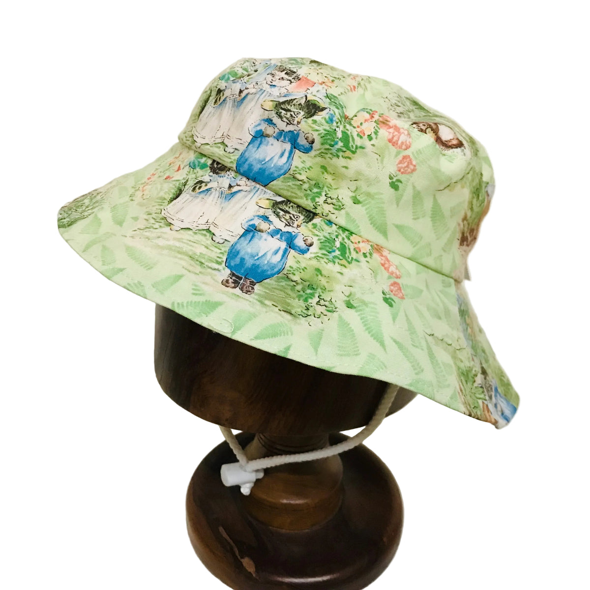 A light green bucket hat with a vintage-style illustration of children in blue dresses and floral patterns, displayed on a dark wooden mannequin stand against a plain off-white background.