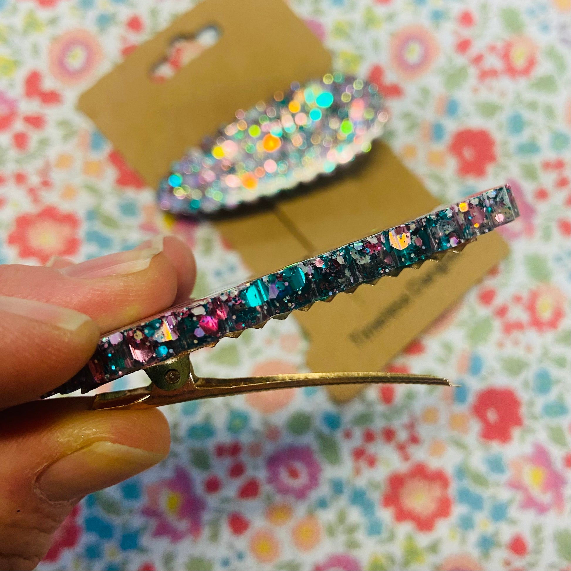 Multicolored glittery hair clip held by a hand with a floral-patterned background