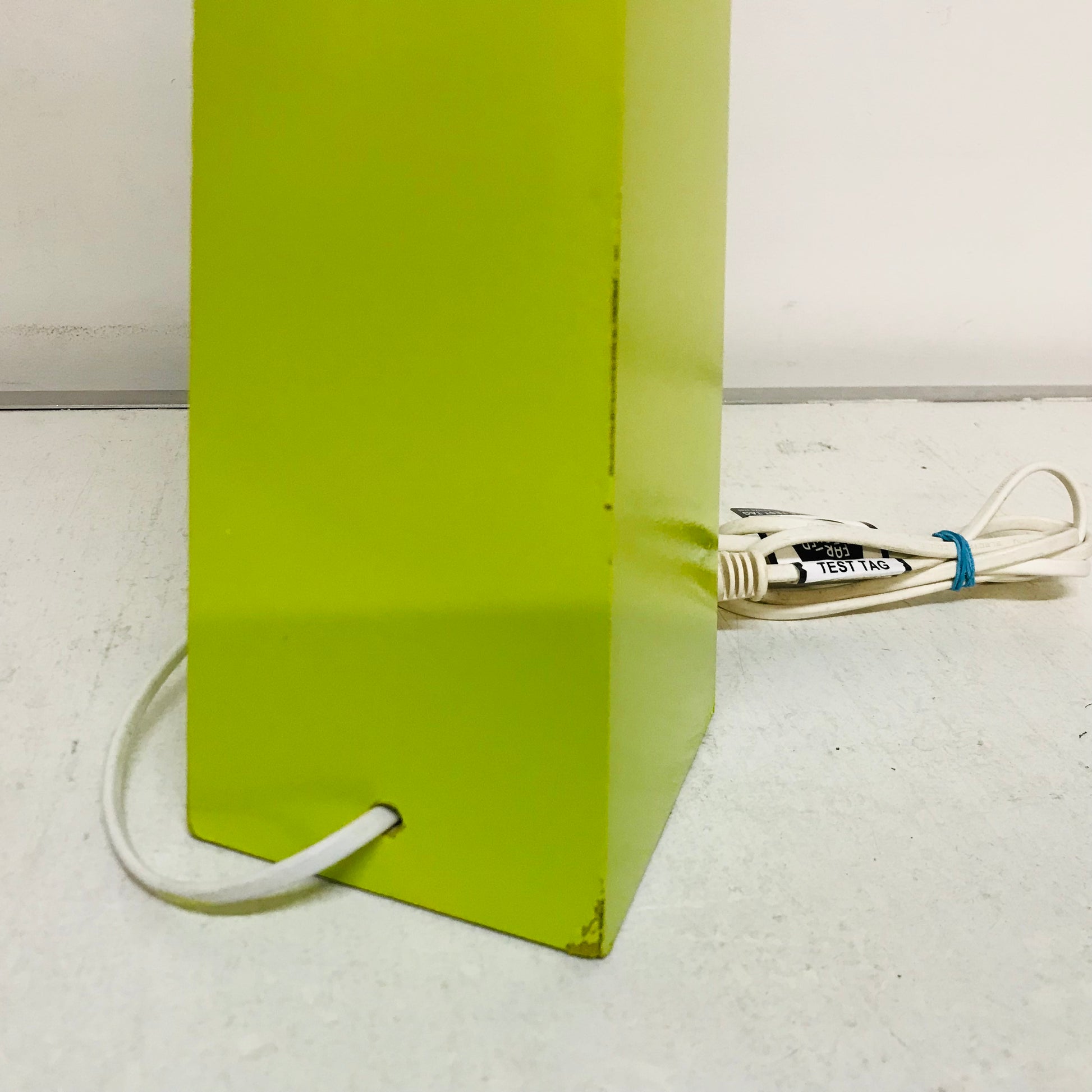 A lime green rectangular metal object, likely a lamp base or speaker, with a white power cord plugged in and looping out the side, set against a plain off-white background.