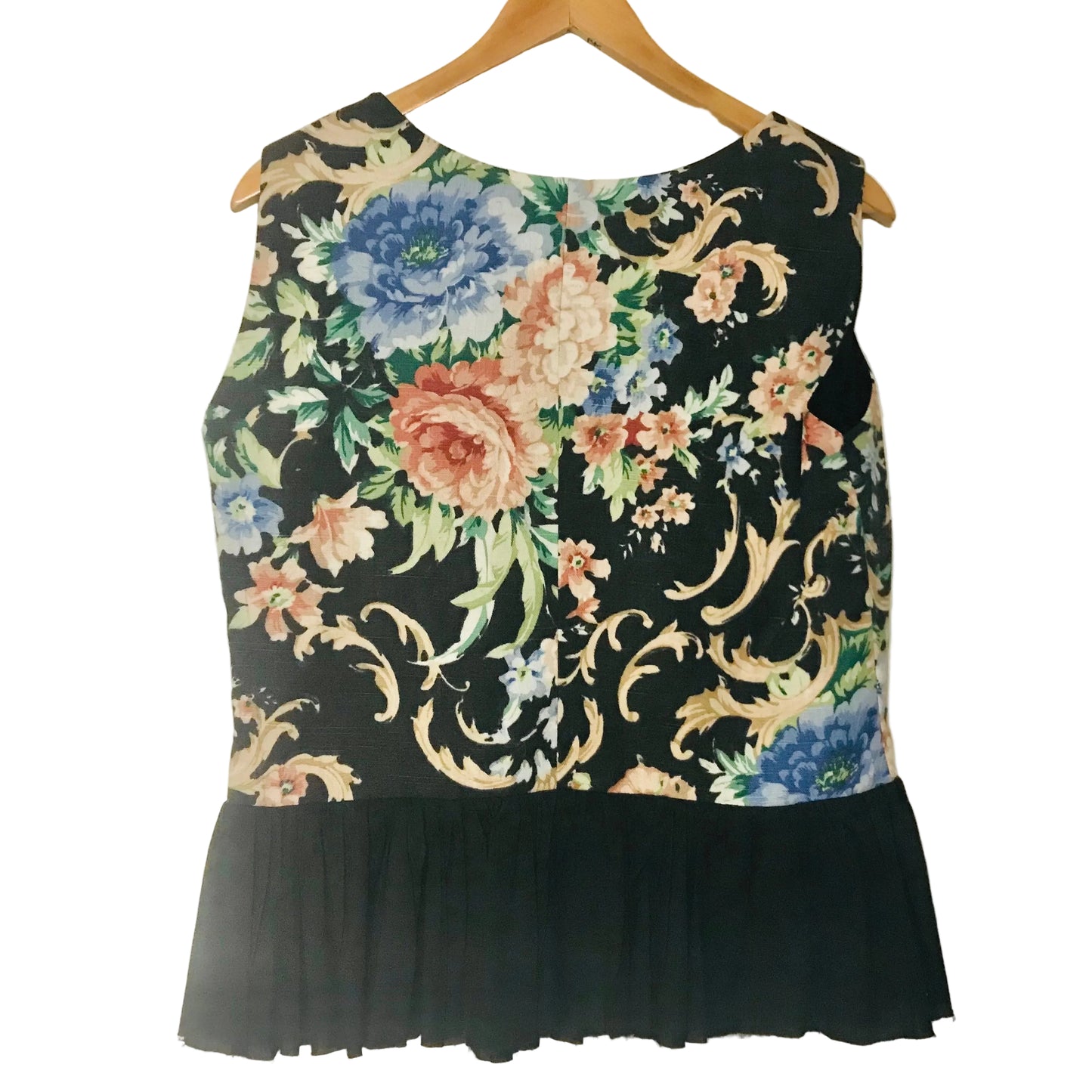 A sleeveless black top with a colorful floral and ornamental print on the upper bodice and a solid black ruffled hem, hanging on a wooden hanger against a plain light wall.