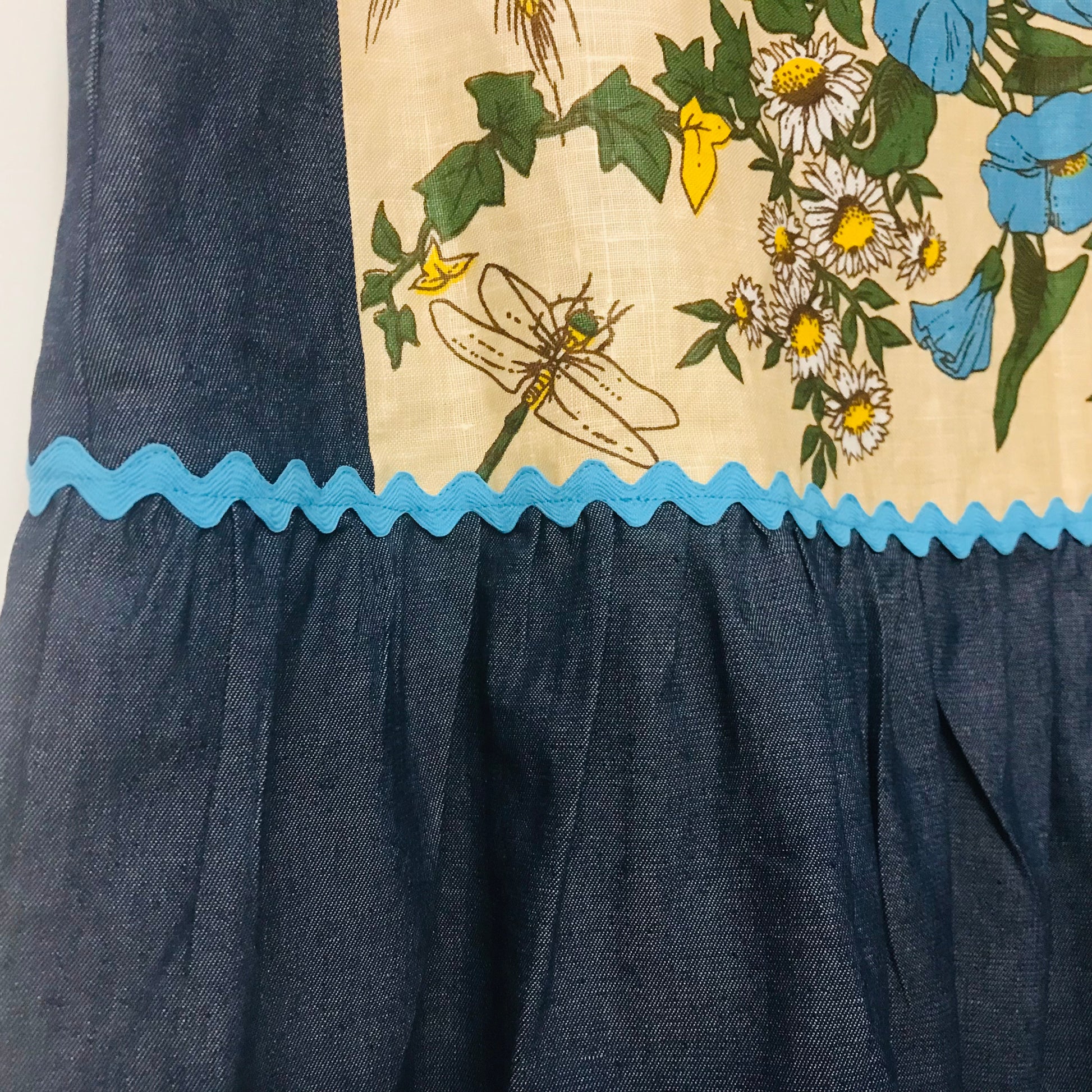Close-up of a garment showing a dark blue textured fabric skirt or lower section meeting a beige printed upper panel with blue flowers, white daisies, green leaves, and a dragonfly, separated by a bright blue scalloped trim.