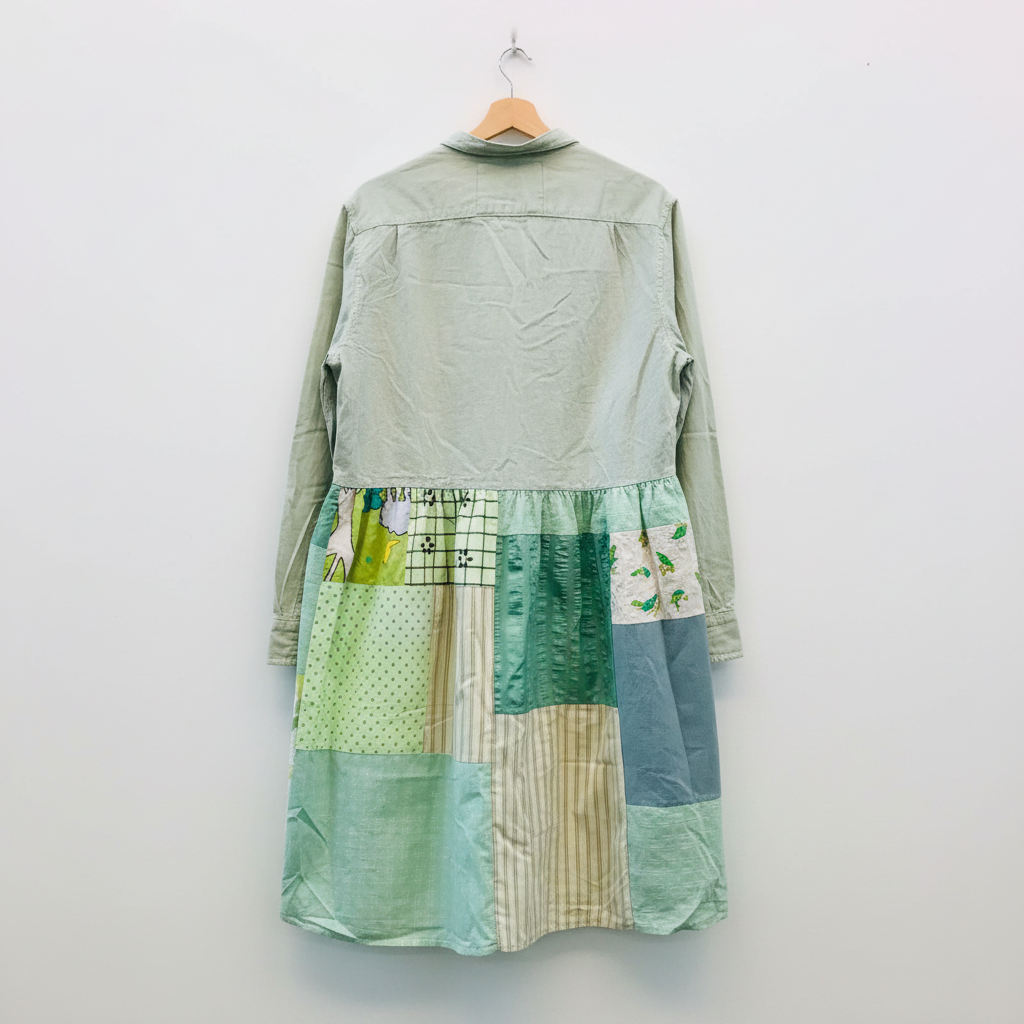 A long-sleeve patchwork dress hanging on a wooden hanger against a plain white wall. The top portion is a solid pale sage green, while the skirt features a patchwork of various green, blue, and beige patterned fabric panels.