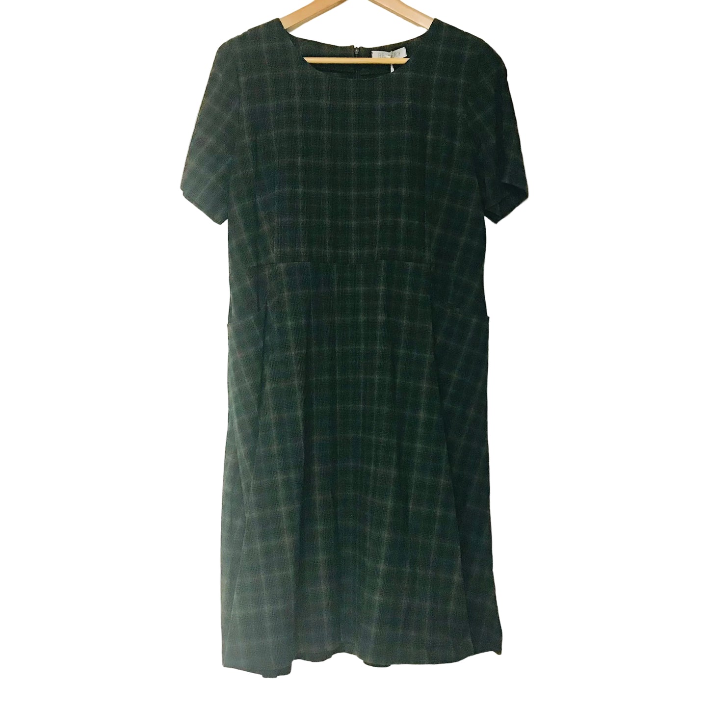 A short-sleeve dark green plaid dress hanging on a wooden hanger against a plain off-white wall.