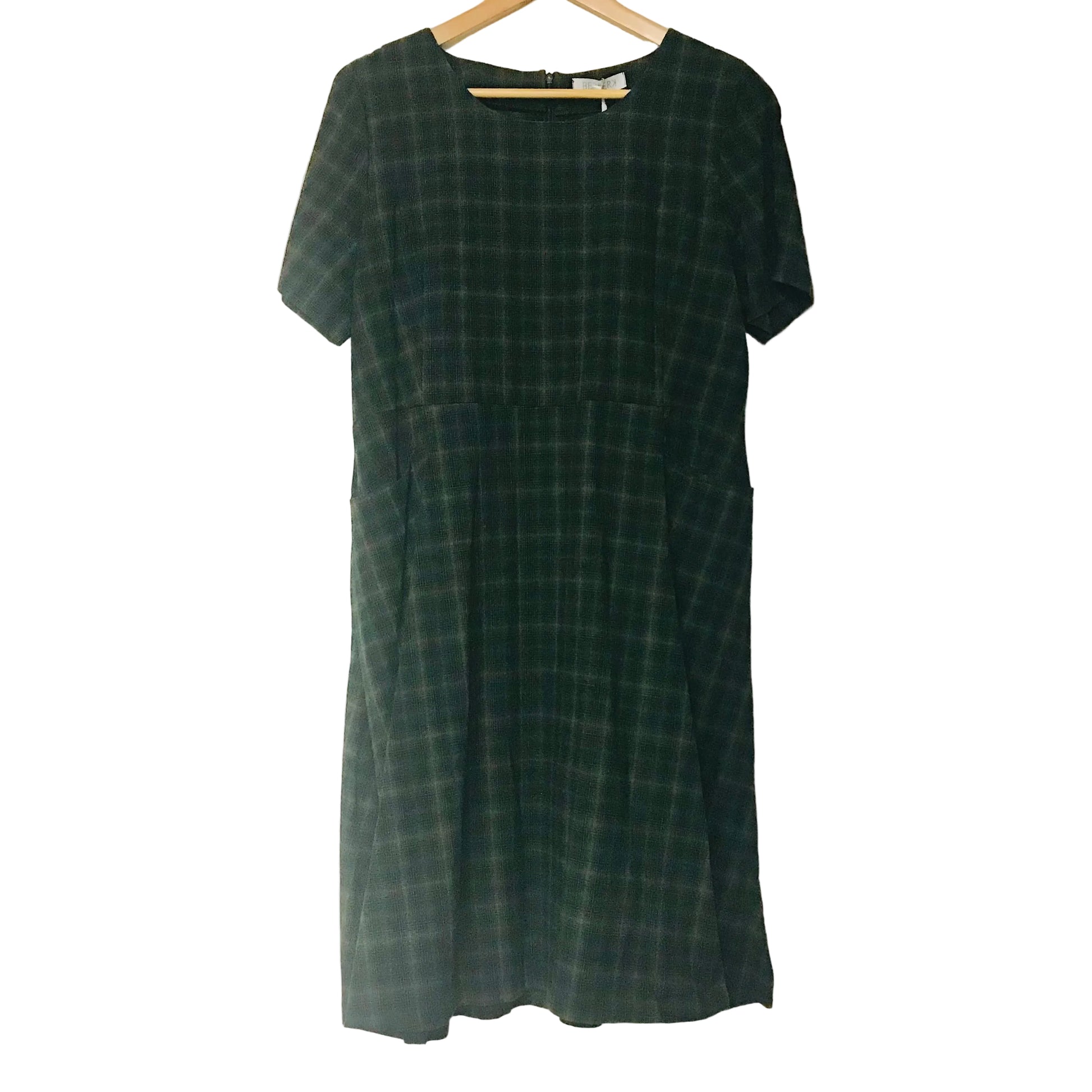 A short-sleeve dark green plaid dress hanging on a wooden hanger against a plain off-white wall.