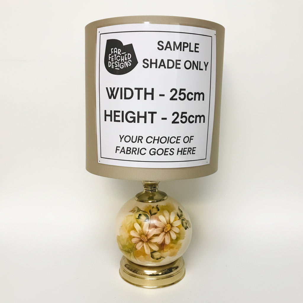 A table lamp shade sample with a large white size and customization label, mounted on a brass neck over a decorative glass globe base with floral patterns.