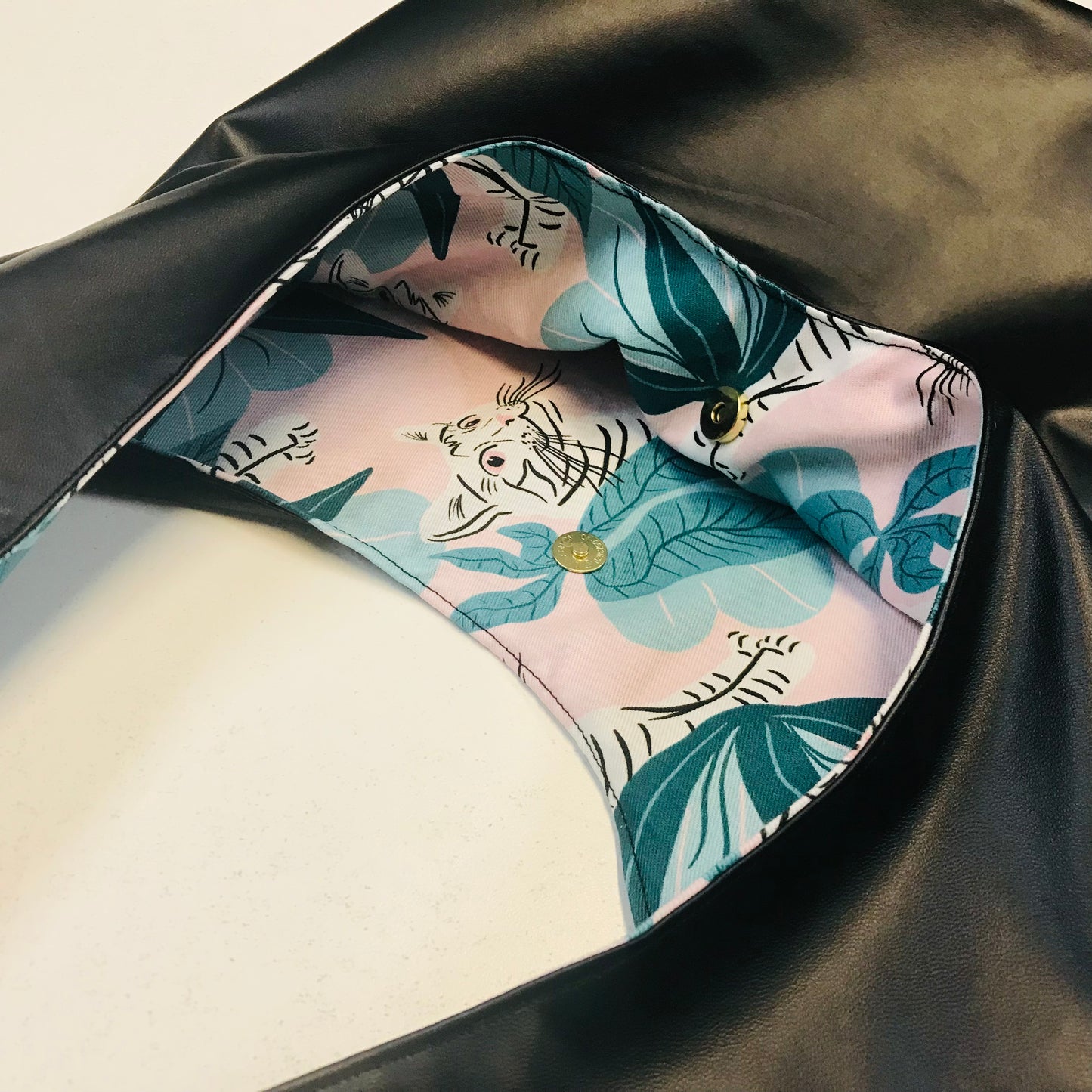 Close-up of a black faux-leather bag interior with a light pink lining printed with teal tropical leaves and a white tiger motif, secured by gold-tone buttons.