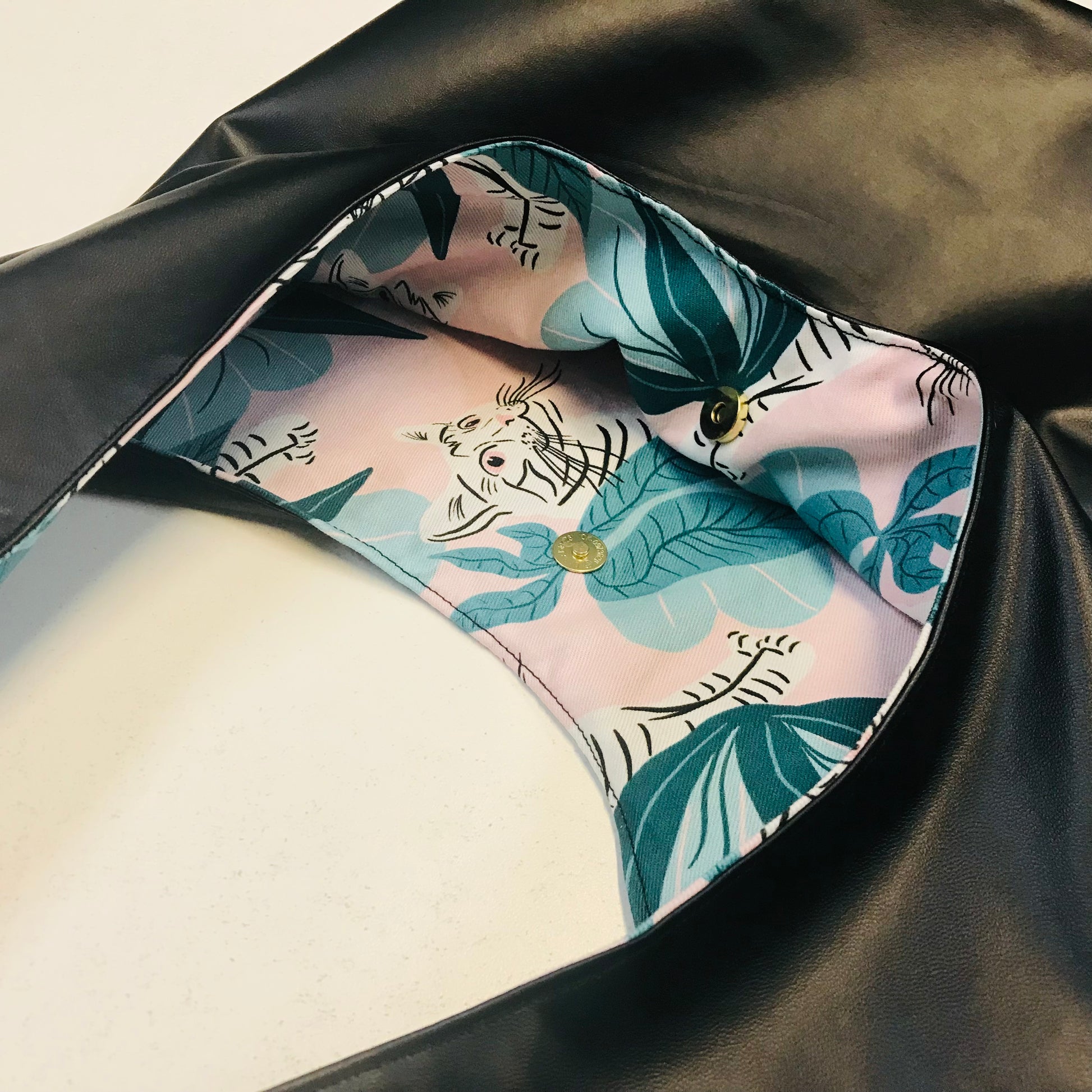 Close-up of a black faux-leather bag interior with a light pink lining printed with teal tropical leaves and a white tiger motif, secured by gold-tone buttons.