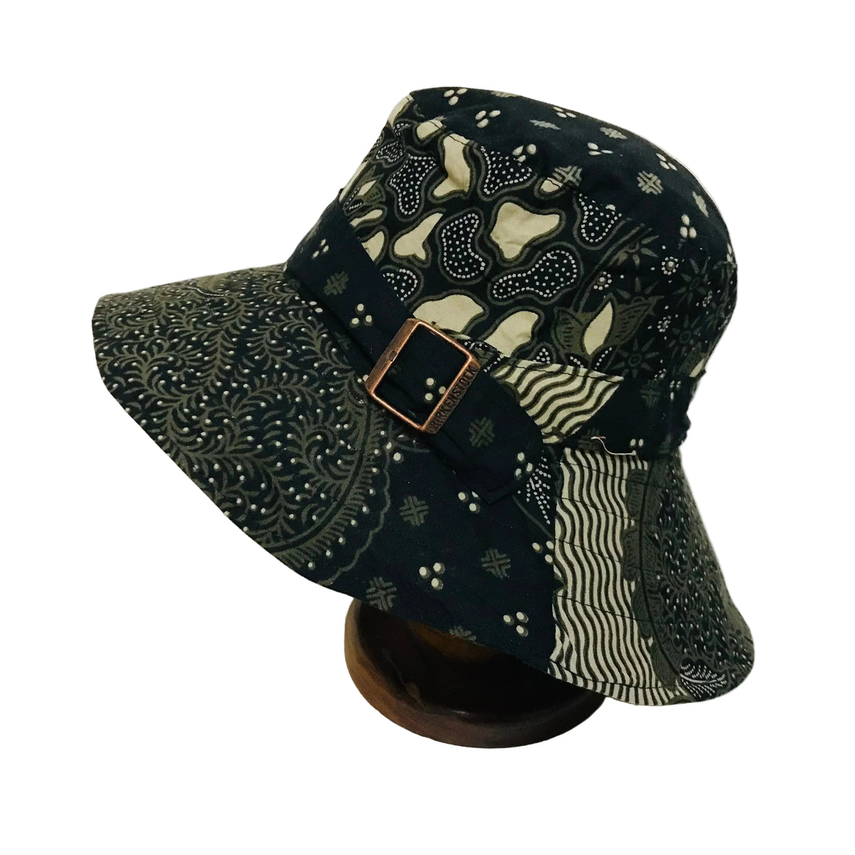 A patchwork bucket hat with a dark green and beige bandana print, wavy stripe pattern, and paisley motif, featuring a brown leather buckle, displayed on a wooden stand against a white background.