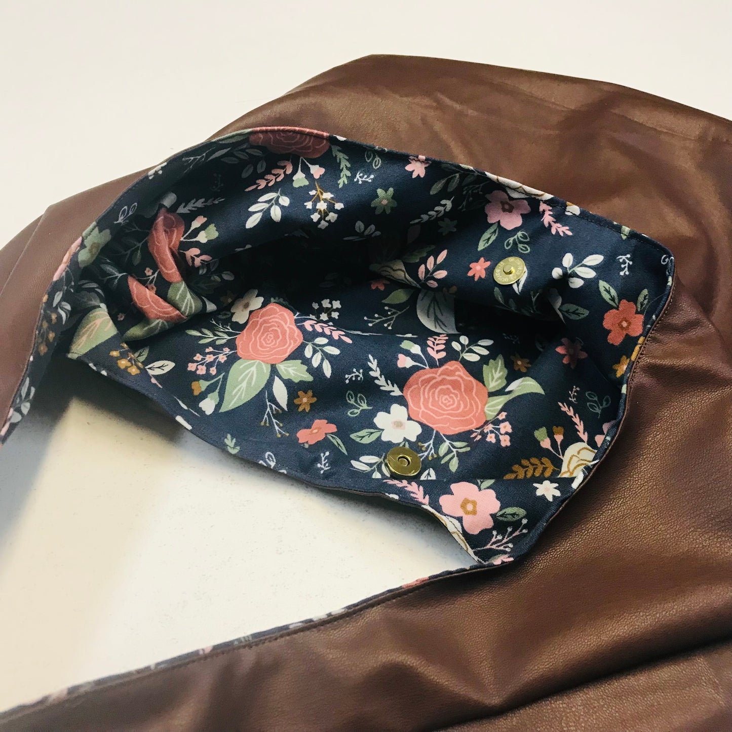 A close-up of a brown faux-leather bag with a floral-patterned fabric lining, photographed on a light neutral background.