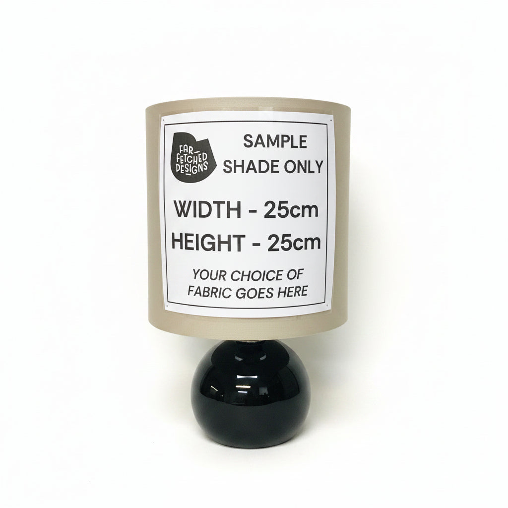 A cylindrical lamp shade mockup with a beige fabric texture and a large white sample label reading 'SAMPLE SHADE ONLY' with dimensions and fabric placeholder text, mounted on a glossy black spherical base against a plain off-white wall.