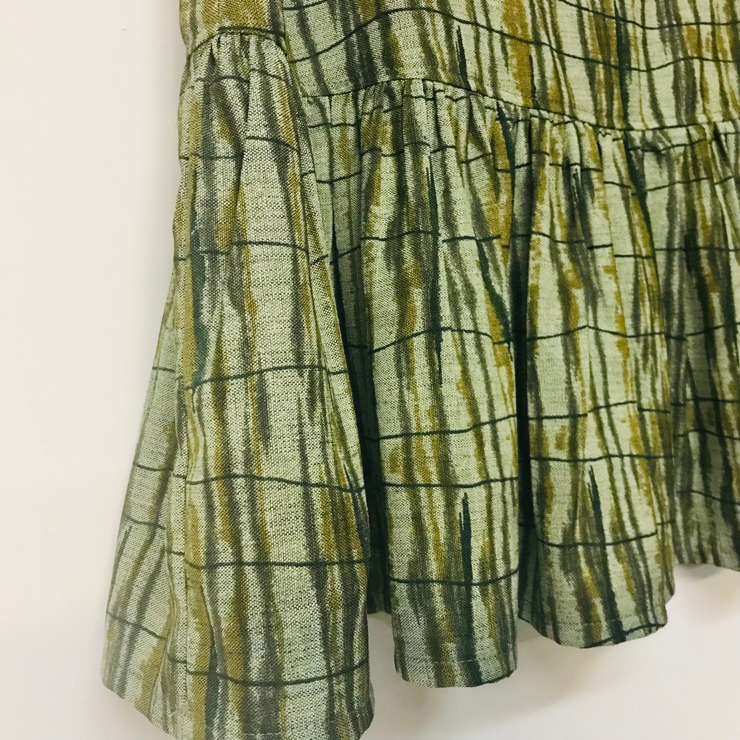 Close-up of a ruffled skirt or dress panel in a textured green and olive fabric with a dark grid plaid pattern, hanging against a plain white wall.