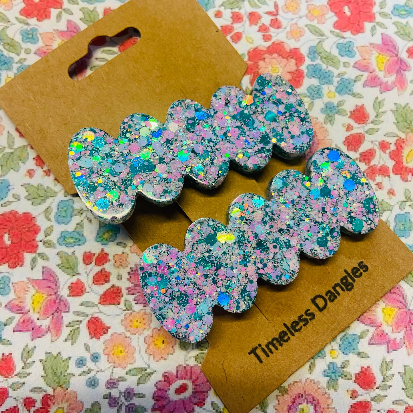 Glittery bow-shaped earrings on a 'Timeless Dangles' card with a floral background