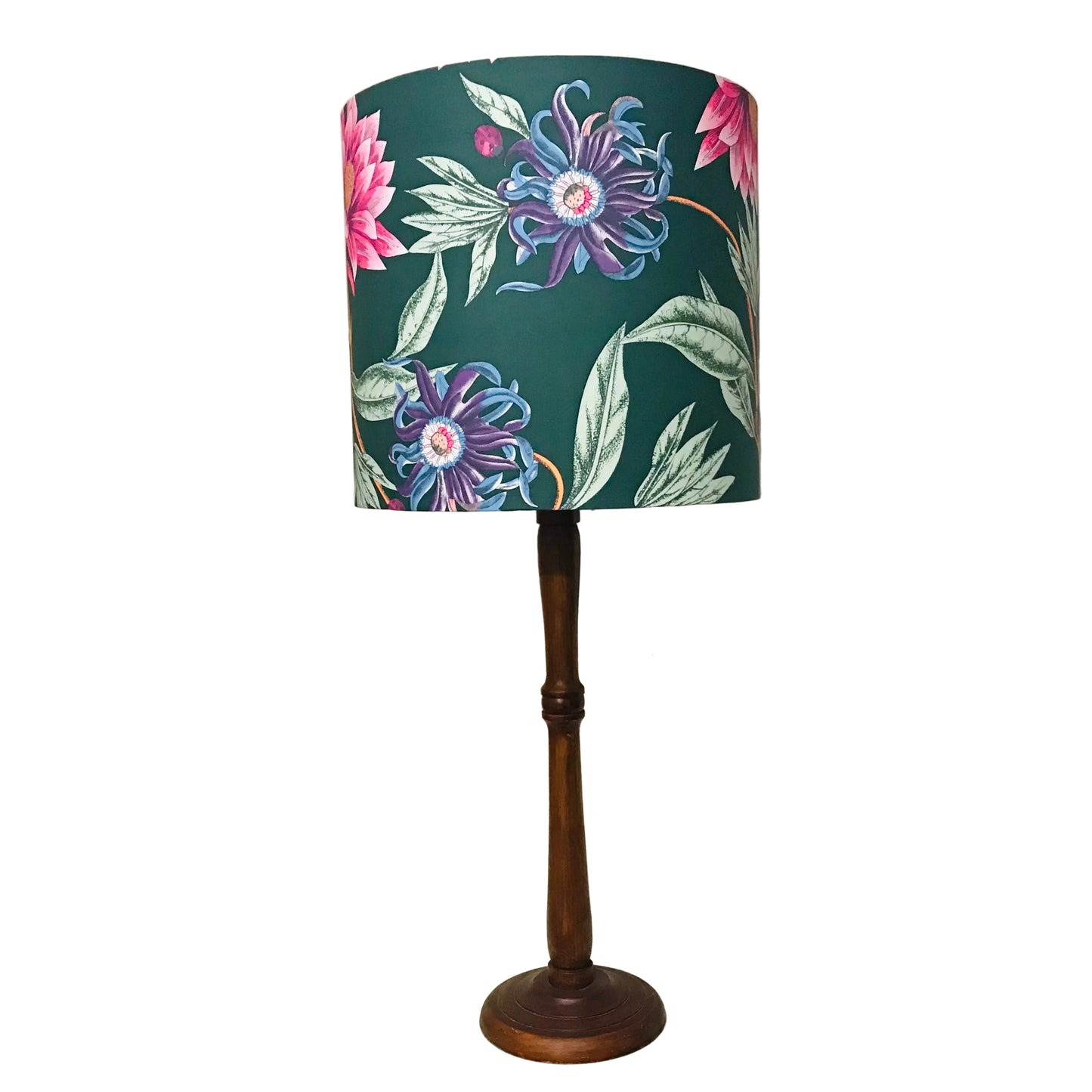A table lamp with a dark green drum shade featuring a vibrant floral print of purple, pink, and blue flowers with green leaves, supported by a dark brown wooden stand, set against a plain white wall.