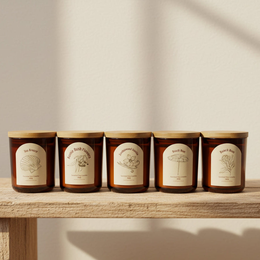 Five amber glass jar candles with bamboo lids are lined up on a light wood table inside a candle shop. The labels feature illustrations and names including 'Sea Breeze', 'Coastal Breeze', 'Woodland Wind', 'Coastal Wind', and 'Sakura Pine'.