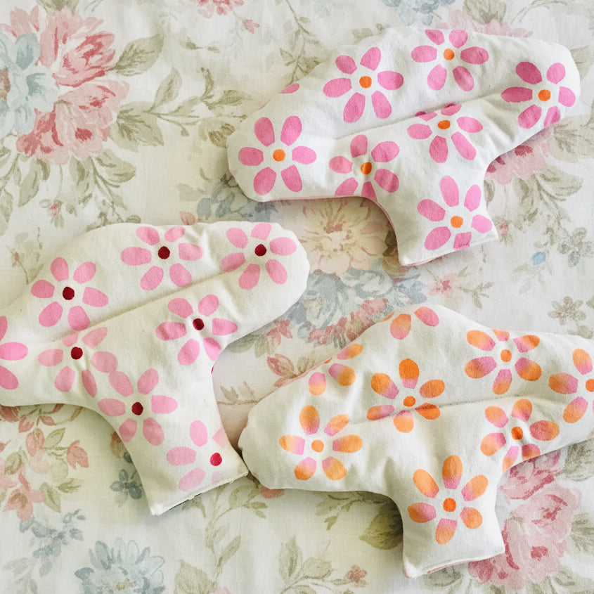 Three reusable menstrual heat pack with a white fabric base and pink and orange floral patterns, arranged on a light wooden surface.