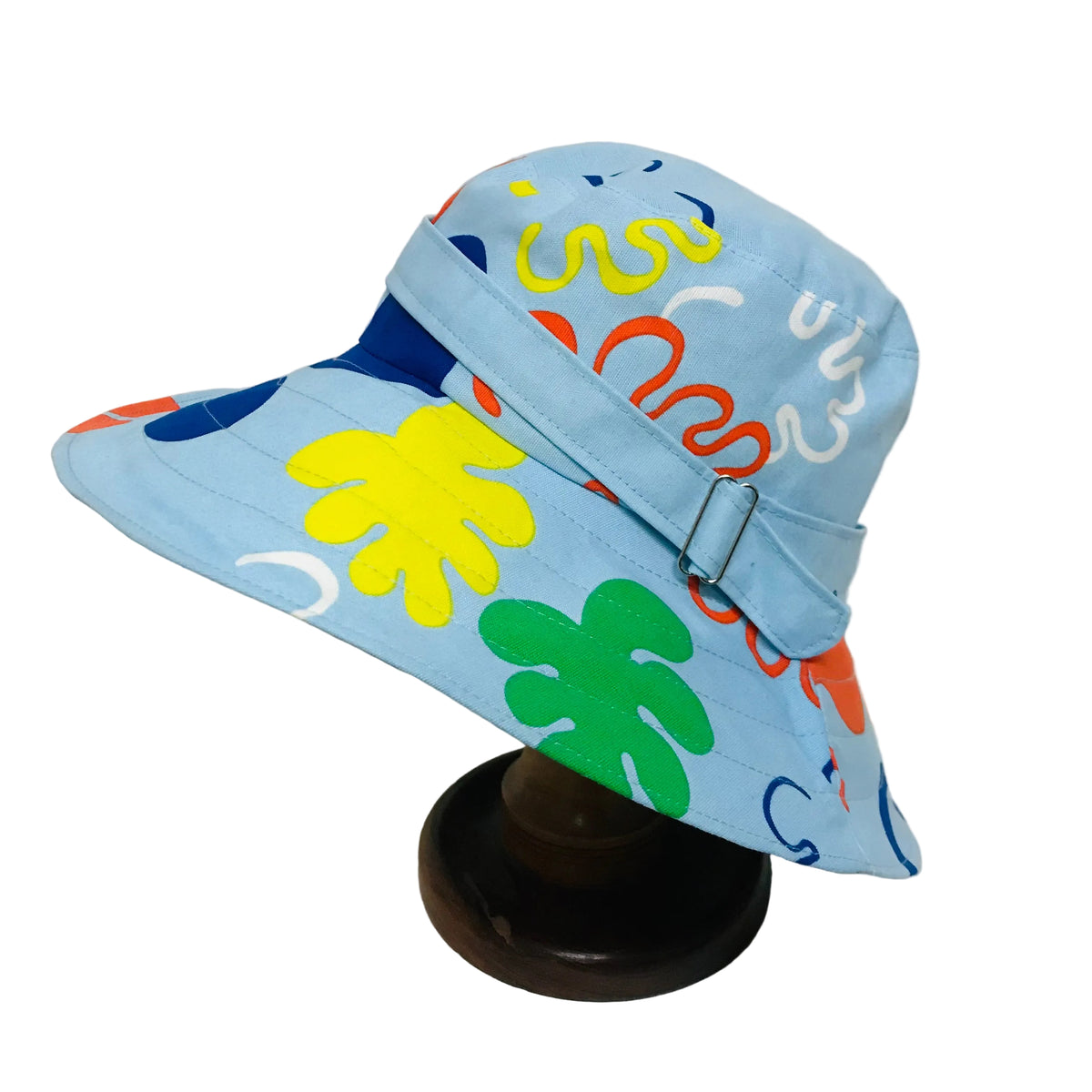A light blue bucket hat with a colorful abstract pattern of wavy lines and tropical leaf shapes, displayed on a dark wooden stand against a plain white background.