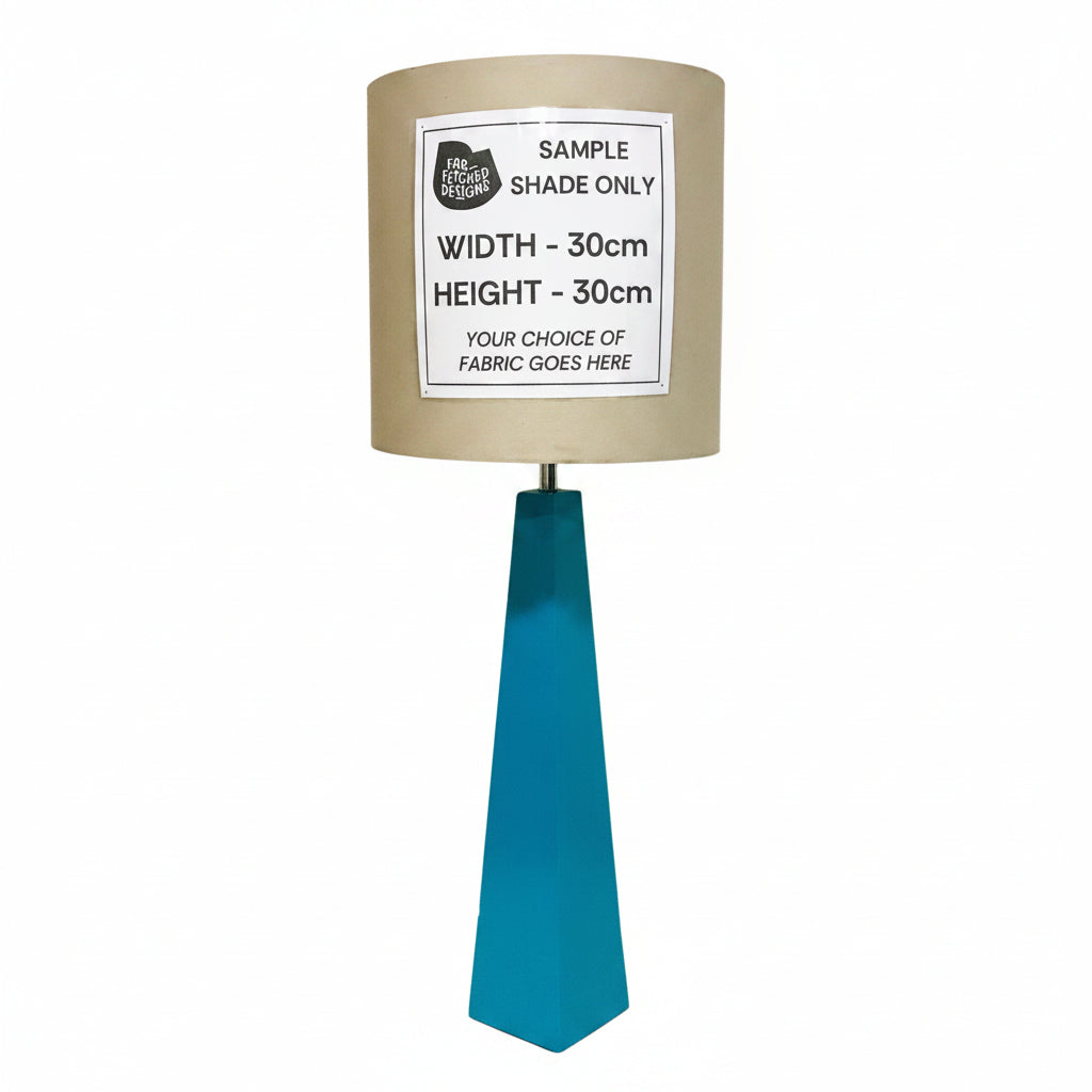 A custom fabric lampshade on a teal geometric metal base, with a white paper label reading 'SAMPLE SHADE ONLY' and dimensions, set against a plain indoor wall.