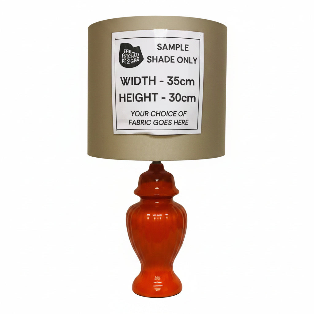 A table lamp with a glossy orange-red fluted base and a beige fabric shade, shown against a plain wall. A white paper label on the shade reads 'SAMPLE SHADE ONLY' with dimensions and fabric placeholder text.