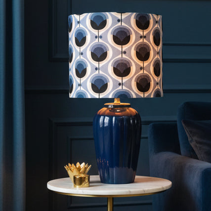 A table lamp with a glossy dark blue ceramic base and a cylindrical drum shade featuring a mid-century modern geometric pattern in shades of blue, navy, cream, and black.