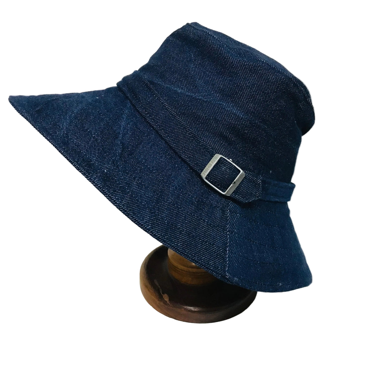A dark blue denim bucket hat with a silver buckle, displayed on a wooden stand against a white background.