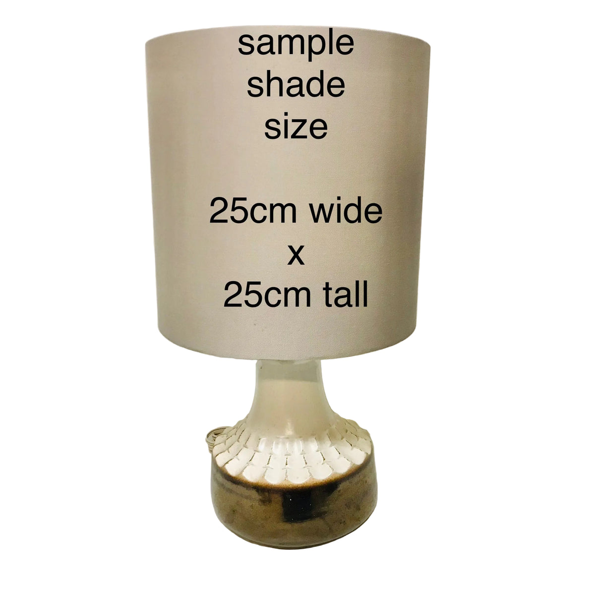 A table lamp shade sample with printed text reading 'sample shade size 25cm wide x 25cm tall' on a beige cylindrical shade, mounted on a decorative glass and ceramic base against a plain wall.