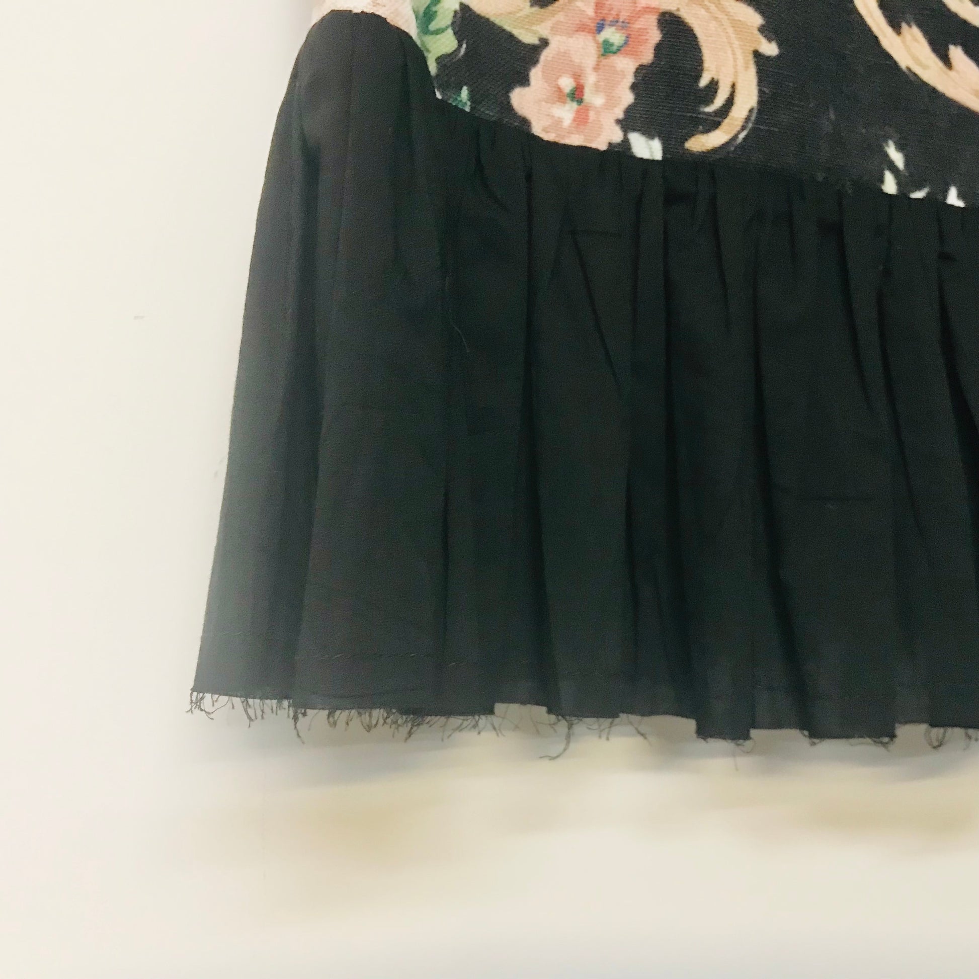 A close-up of a black pleated skirt or drape with raw frayed edges, shown against a plain light background with a floral pattern partially visible at the top.