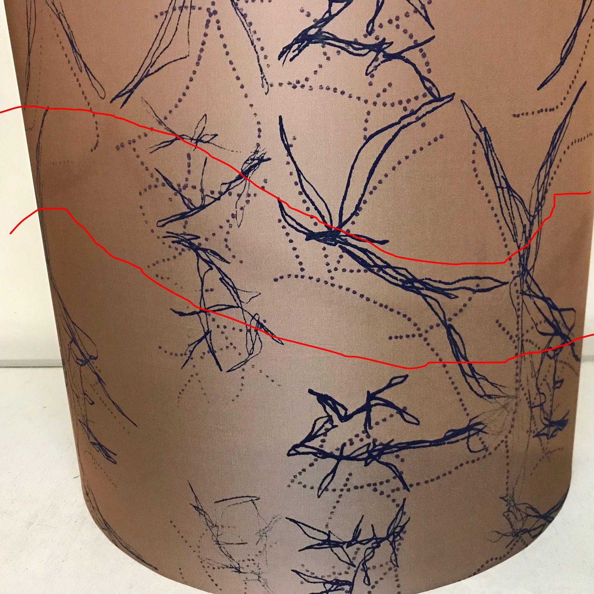 A close-up of a cylindrical lampshade or drum shade in warm taupe-beige with a dark navy abstract line and dotted pattern across the surface.