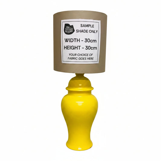 A yellow ceramic table lamp with a brown fabric shade, featuring a white sample label on the shade that reads 'SAMPLE SHADE ONLY' with dimensions and placeholder text.