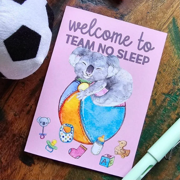 greeting card with a koala illustration and text 'Welcome to Team No Sleep' on a wooden surface.