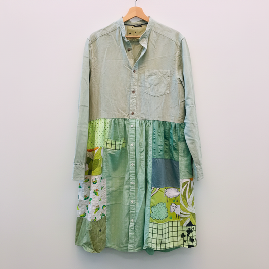 A long-sleeve patchwork shirt dress hanging on a wooden hanger against a white brick wall. The top portion is a muted sage green linen-like button-up, while the lower skirt section features various green and yellow patchwork panels with floral and grid patterns.