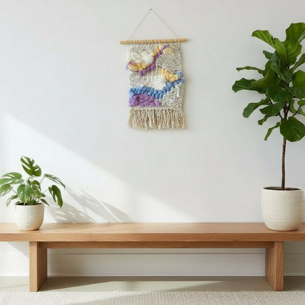 Wooden bench with two potted plants and a colorful wall hanging against a white wall.