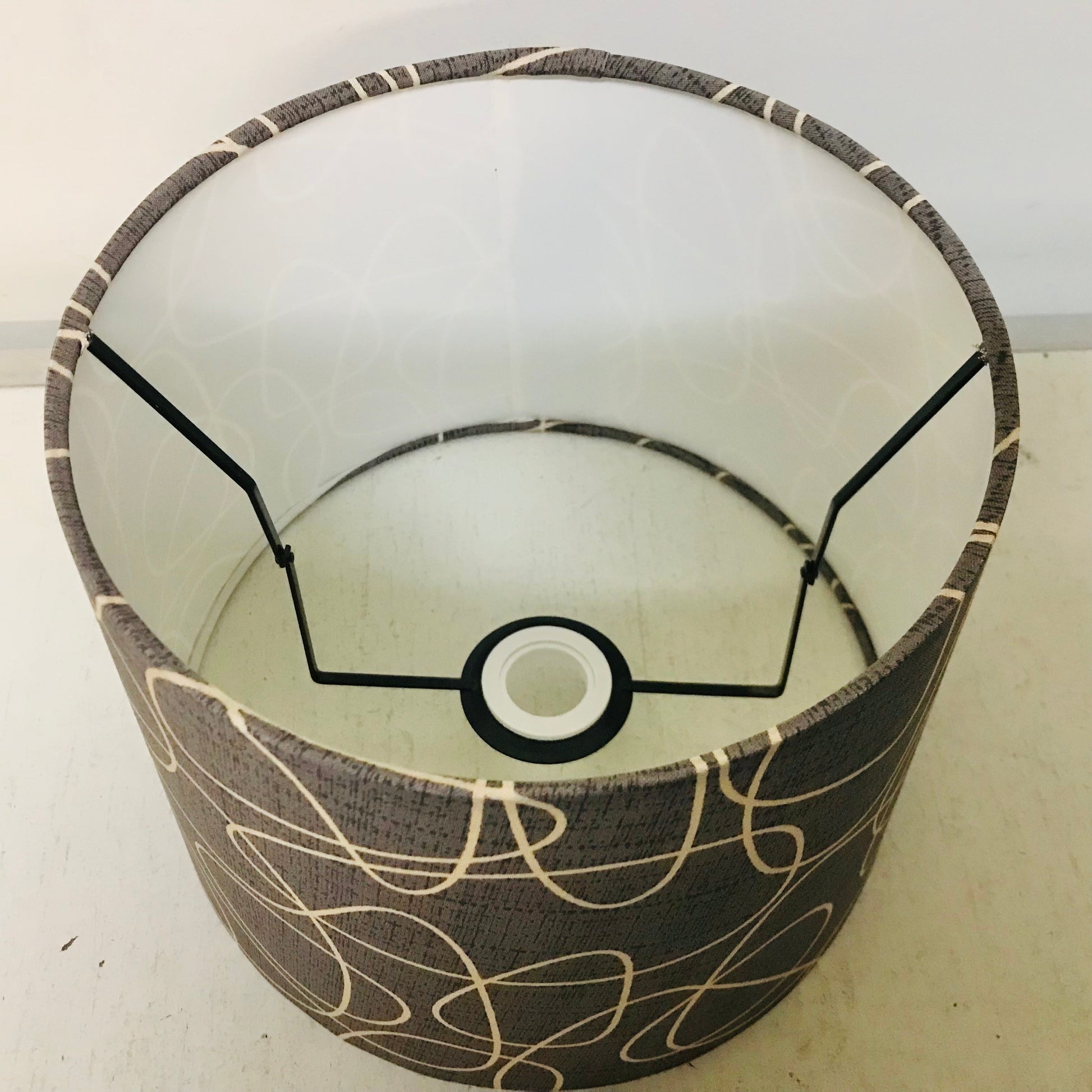 A cylindrical fabric lampshade with a dark gray textured base and beige abstract line patterns, shown with its black metal support frame inside.