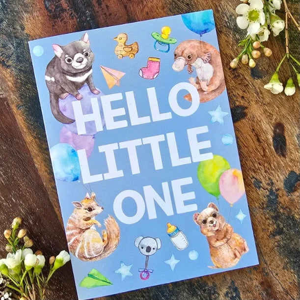 Card with cartoon animals and 'Hello Little One' text on a wooden surface with flowers.