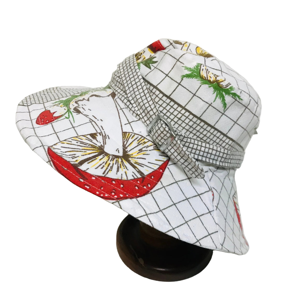 A white bucket hat with a black grid pattern and colorful illustrations of strawberries, green leaves, and yellow flowers, displayed on a dark wooden stand against a plain off-white background.