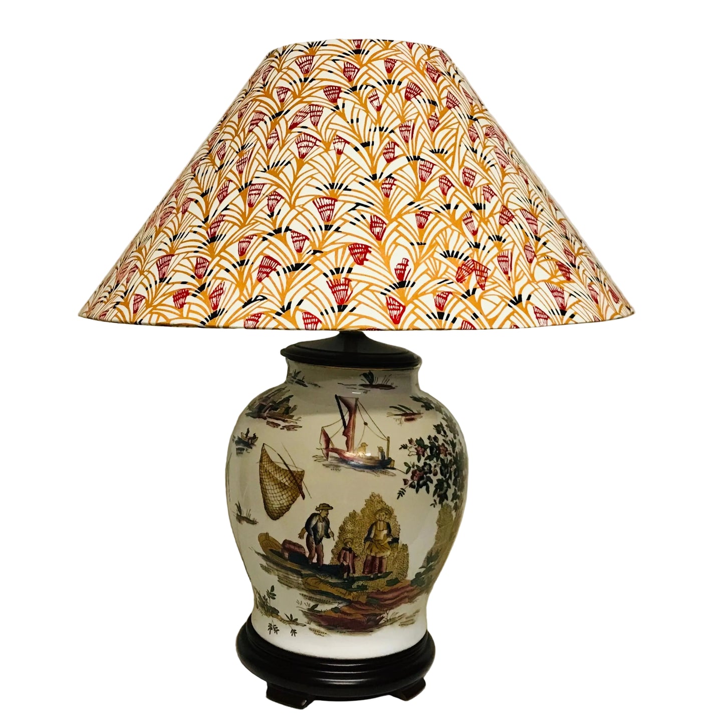 A table lamp with a cream-colored ceramic base decorated with a scenic landscape print, featuring a large conical shade with a repeating orange, red, and black fan pattern, set against a plain off-white wall.