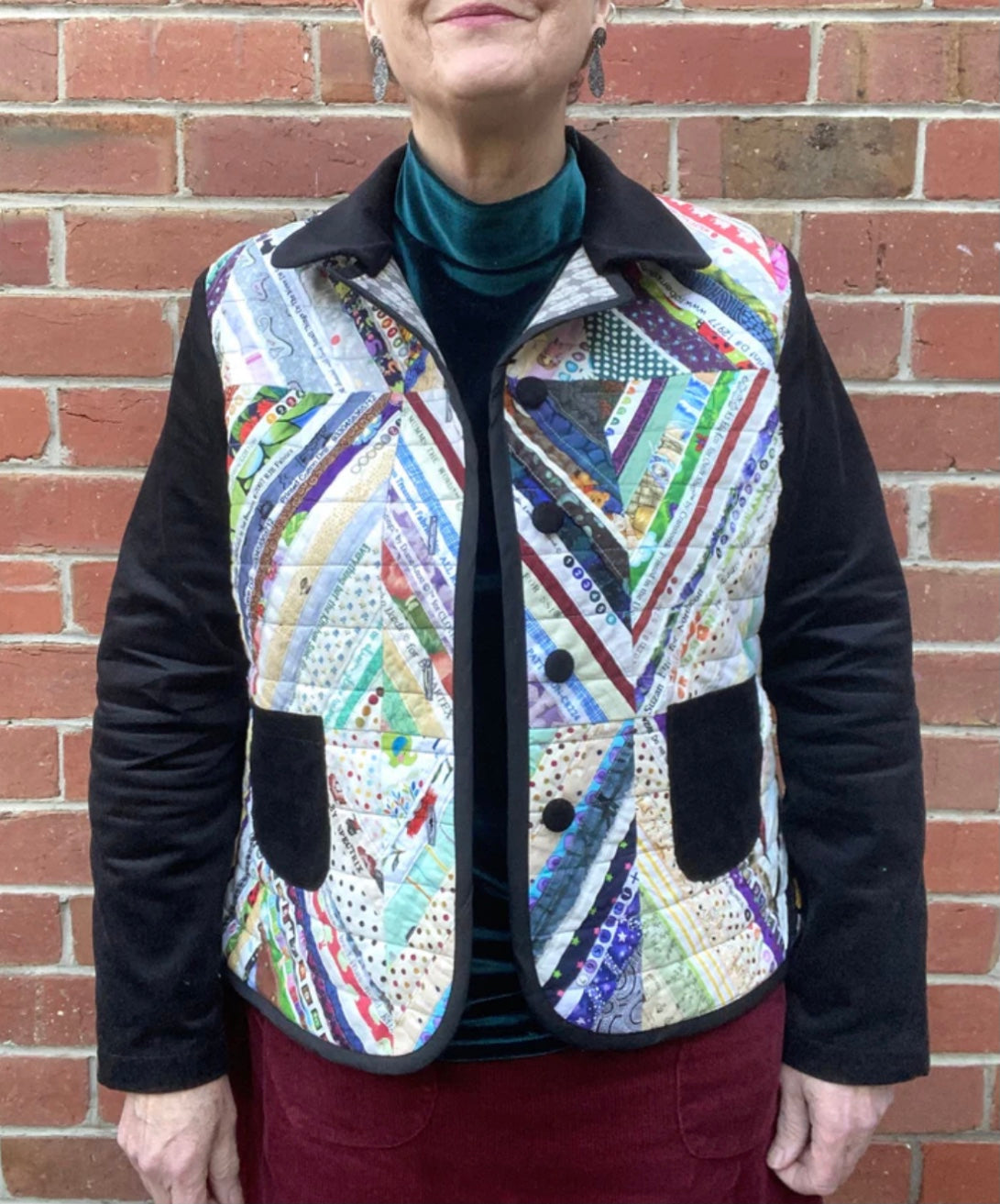 Person wearing a colorful quilted jacket with black sleeves against a brick wall.