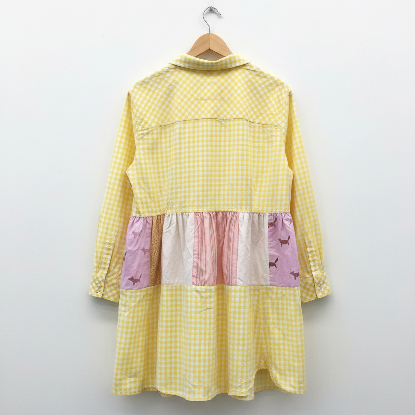 A yellow and white gingham patchwork dress hanging on a wooden hanger against a white brick wall. The dress features a solid light pink ruffled band across the waist and matching pink and lavender patchwork details on the sleeves and lower skirt.