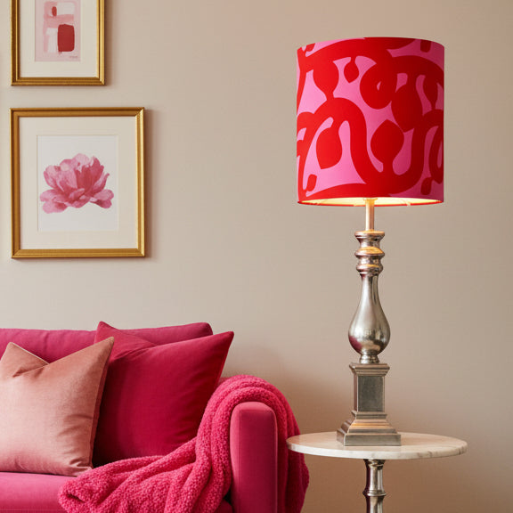 A table lamp with a cylindrical drum shade featuring a bold red and pink abstract pattern, mounted on a polished silver metal base.