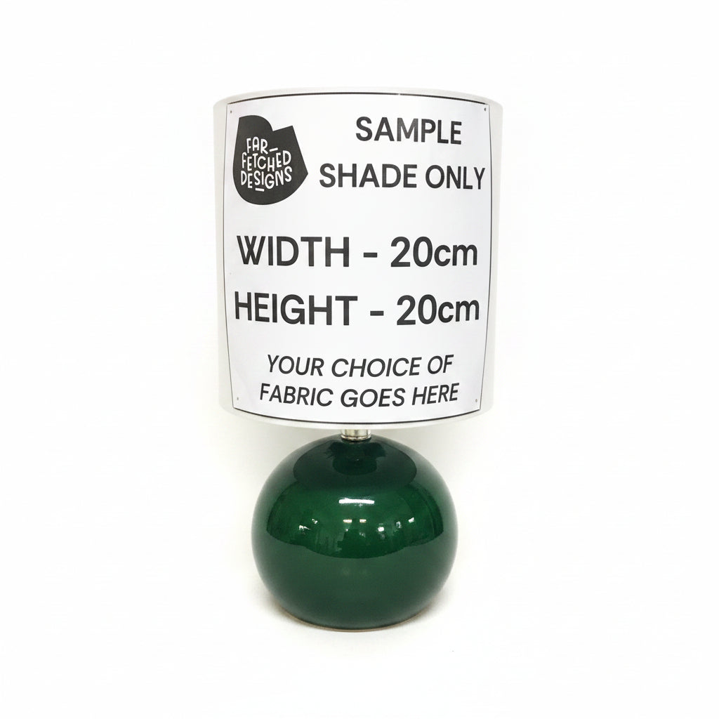 A black spherical table lamp with a beige drum shade, featuring a white paper label that reads 'SAMPLE SHADE ONLY' and dimensions 'WIDTH - 20cm HEIGHT - 20cm'.